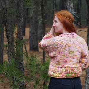 May include: A pink, yellow, and green knit sweater with a textured design. The sweater has a round neckline and long sleeves. The model is wearing the sweater in a forest setting.