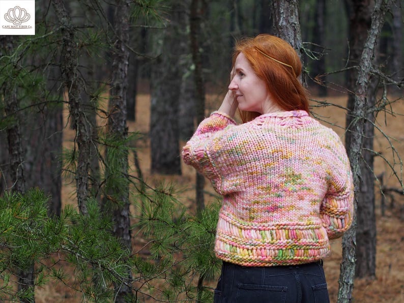 May include: A pink, yellow, and green knit sweater with a textured design. The sweater has a round neckline and long sleeves. The model is wearing the sweater in a forest setting.