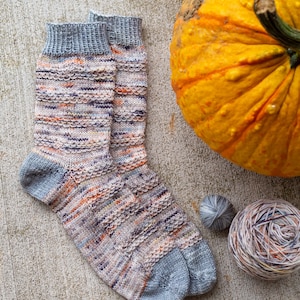 May include: A pair of hand-knitted socks in a multi-colored yarn with a grey heel and toe. The socks are lying on a concrete surface next to a pumpkin and two balls of yarn.