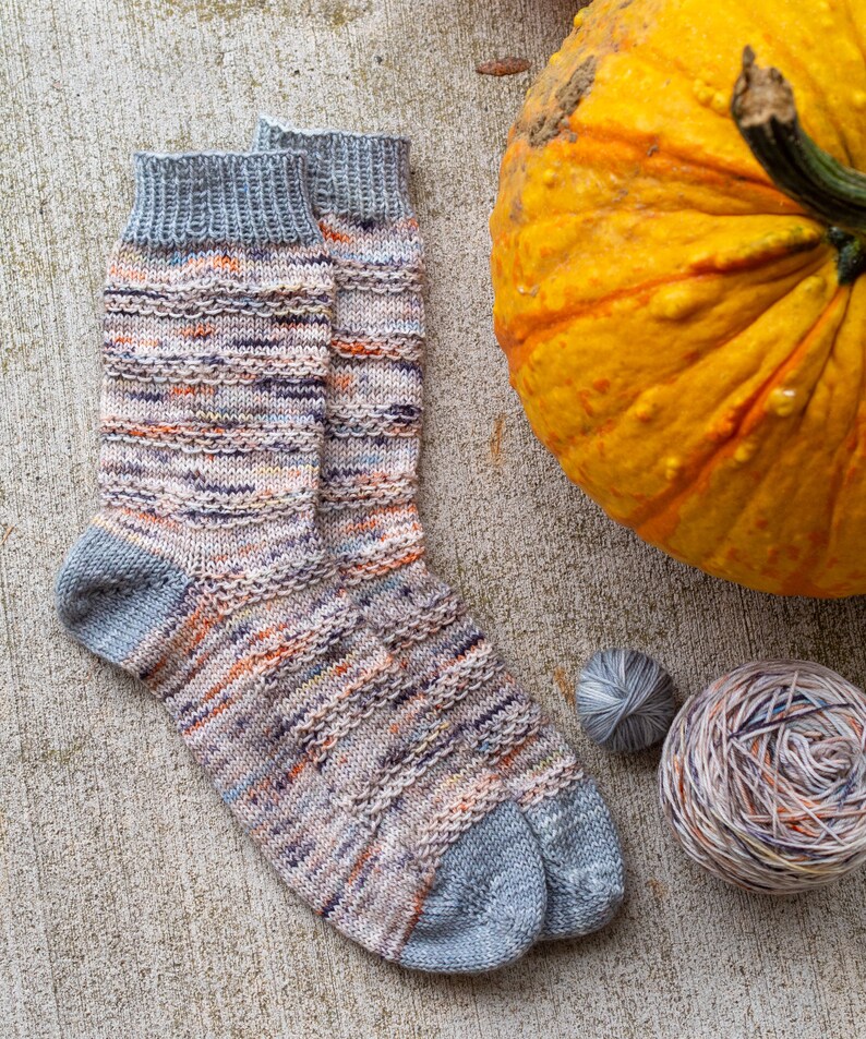 May include: A pair of hand-knitted socks in a multi-colored yarn with a grey heel and toe. The socks are lying on a concrete surface next to a pumpkin and two balls of yarn.