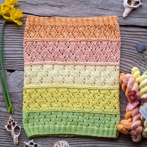 May include: A multi-colored knitted cowl in shades of orange, pink, yellow, and green. The cowl features intricate lace patterns and is displayed with yarn skeins, seashells, and daffodils. The logo "Cape May Fiber Co." is visible.
