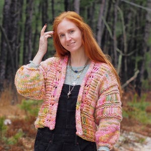 May include: A hand-knitted, open-front cardigan in shades of pink, yellow, and green. The chunky knit sweater has a textured appearance with a relaxed fit and long sleeves. The model is wearing black overalls. The Cape May Fiber Co. logo is in the upper left corner.