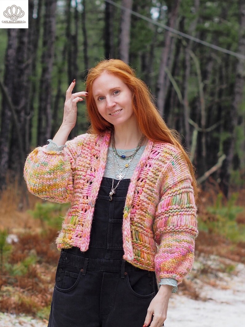 May include: A hand-knitted, open-front cardigan in shades of pink, yellow, and green. The chunky knit sweater has a textured appearance with a relaxed fit and long sleeves. The model is wearing black overalls. The Cape May Fiber Co. logo is in the upper left corner.
