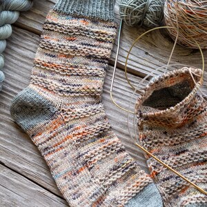 May include: A pair of hand-knitted socks in a multi-colored yarn with a grey heel and toe. The socks are lying on a wooden surface with yarn and knitting needles.