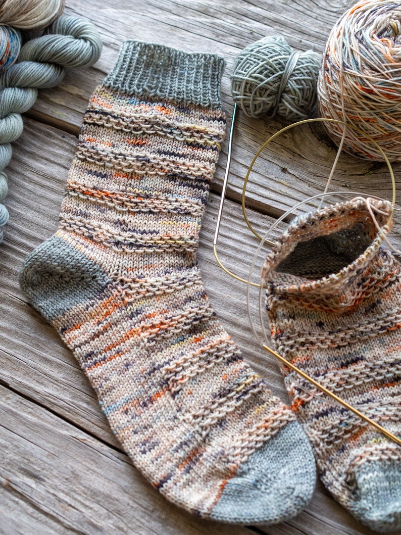 May include: A pair of hand-knitted socks in a multi-colored yarn with a grey heel and toe. The socks are lying on a wooden surface with yarn and knitting needles.