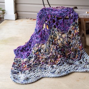 May include: A large, hand-knitted blanket in shades of purple, blue, and grey with a lacy pattern. The blanket is draped over a wooden bench.