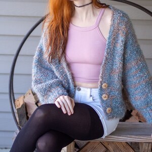 May include: A woman wearing a light blue and brown fuzzy cardigan over a pink tank top and white denim shorts. She is sitting on a wooden bench with her legs crossed and wearing black tights.