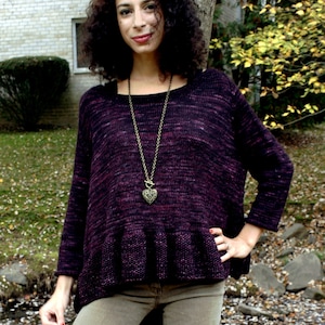 May include: A woman wearing a purple and black knit sweater with a ruffled hem and a long gold chain necklace with a heart pendant. She is wearing brown pants and is standing in front of a tree.