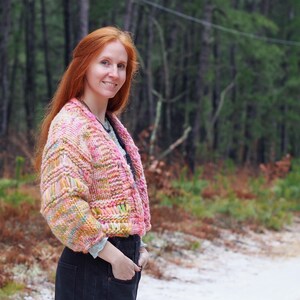 May include: A cropped, chunky knit cardigan in shades of pink, yellow, and green. The open-front sweater has a textured pattern and is worn over a gray shirt and black jeans. The background features a forest and a snowy road.