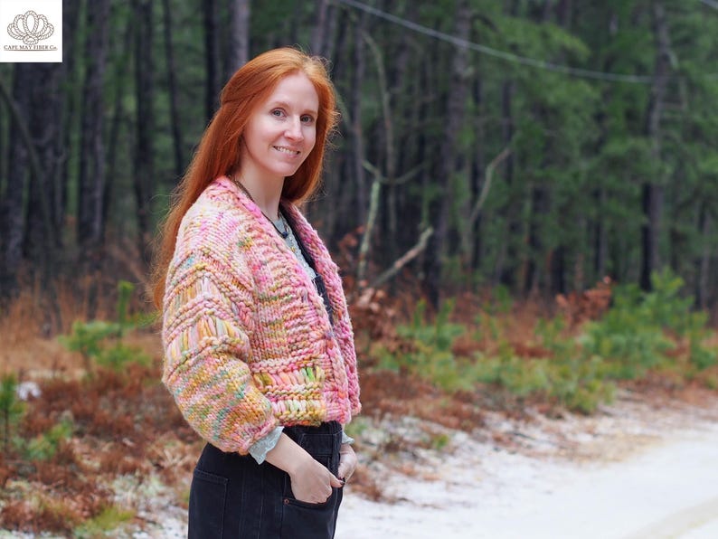 May include: A cropped, chunky knit cardigan in shades of pink, yellow, and green. The open-front sweater has a textured pattern and is worn over a gray shirt and black jeans. The background features a forest and a snowy road.