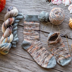 May include: A pair of hand-knitted socks in a multi-colored yarn with a grey heel and toe. The socks are lying on a wooden surface with yarn balls, seashells, and pumpkins.
