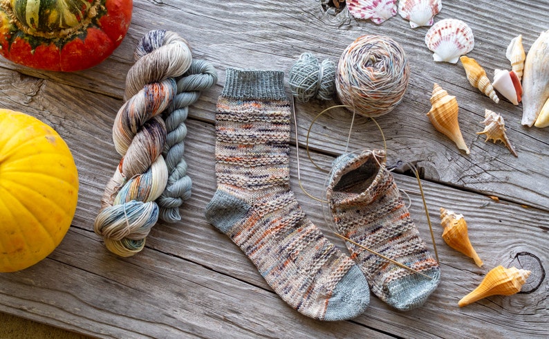 May include: A pair of hand-knitted socks in a multi-colored yarn with a grey heel and toe. The socks are lying on a wooden surface with yarn balls, seashells, and pumpkins.