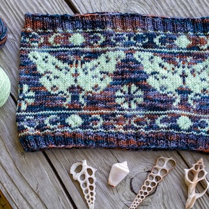 May include: A knitted headband with a colorful pattern of butterflies and snowflakes. The headband is made with a variety of shades of blue, green, brown, and white yarn.