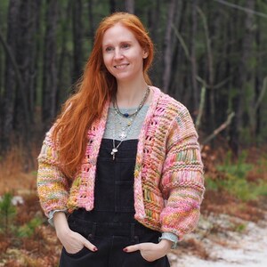 May include: A woman wearing a colorful, chunky knit cardigan with pink, yellow, and green hues. The cropped sweater has a textured design and is paired with black overalls and layered necklaces. The background features a blurred forest.
