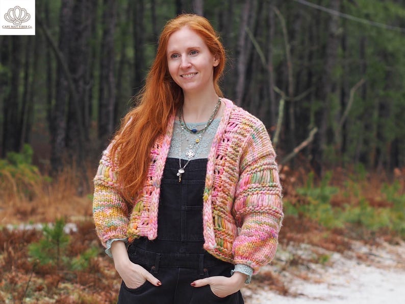 May include: A woman wearing a colorful, chunky knit cardigan with pink, yellow, and green hues. The cropped sweater has a textured design and is paired with black overalls and layered necklaces. The background features a blurred forest.