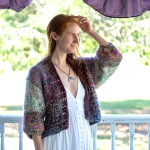 May include: A woman wearing a white dress with a multi-colored knitted cardigan. The cardigan has a loose fit and a cropped length. The woman is standing on a porch with a white railing.