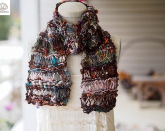 May include: A handcrafted scarf with a unique, textured design. The scarf features a mix of brown, blue, and pink yarns, creating a visually rich and intricate pattern. The scarf is displayed on a mannequin, showcasing its shape and drape.