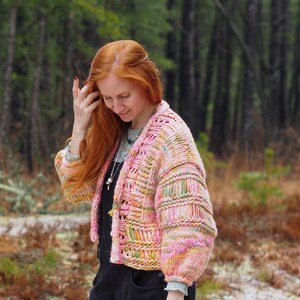 May include: A colorful, hand-knitted cardigan in shades of pink, yellow, and green. The open-front sweater features a textured stitch pattern and voluminous sleeves. The image is set outdoors with a blurred forest background.