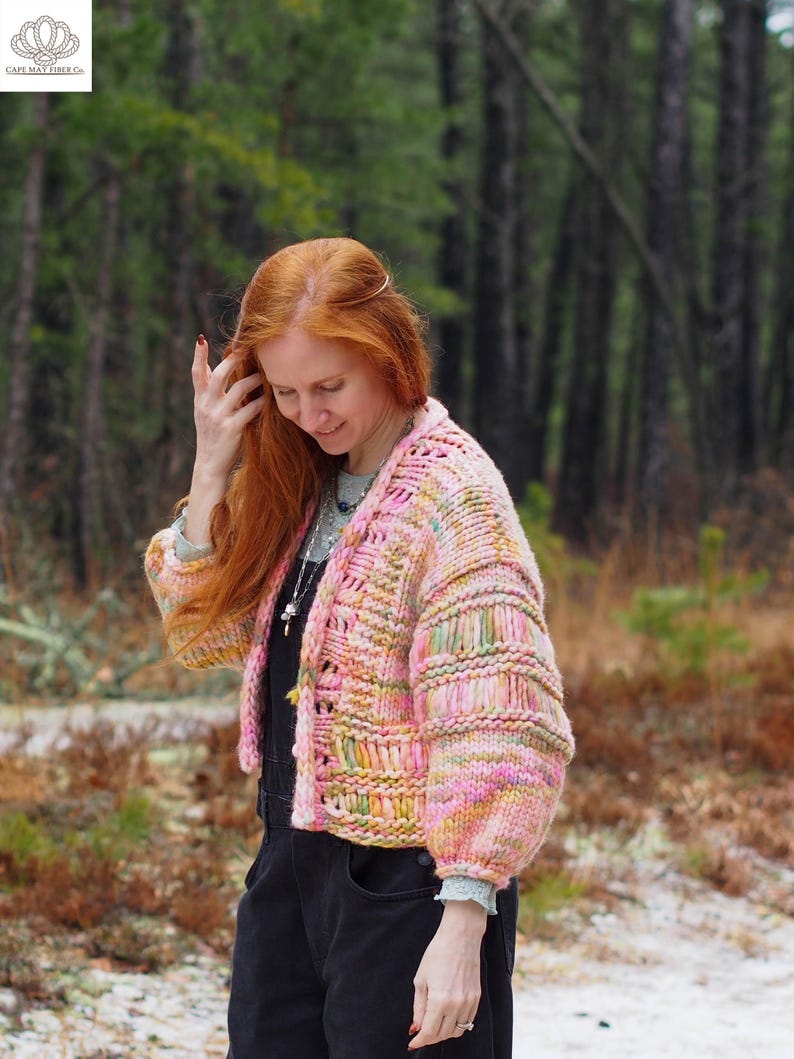 May include: A colorful, hand-knitted cardigan in shades of pink, yellow, and green. The open-front sweater features a textured stitch pattern and voluminous sleeves. The image is set outdoors with a blurred forest background.