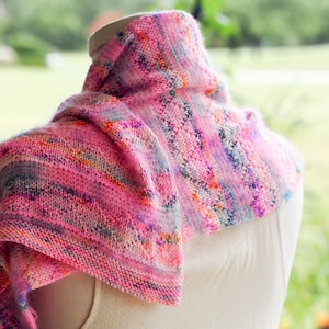 May include: A hand-knitted cowl scarf in shades of pink, purple, and orange. The scarf is draped over a mannequin, showcasing its textured knit and color variations. The yarn appears soft and warm, ideal for cooler weather.