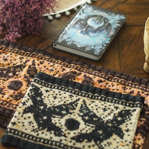 Puede incluir: Primer plano de dos artículos de punto con intrincados patrones en colores marrón, naranja y crema. También se ve un pequeño cuaderno con un diseño celestial y una ramita de flores moradas. Los artículos parecen hechos a mano.