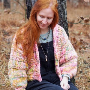 May include: A chunky knit cardigan in pastel shades of pink, yellow, and green. The open-front sweater has a boxy silhouette with wide sleeves. The garment is handmade with a textured stitch, and the sleeves are cuffed. The cardigan is worn over a dark jumpsuit.