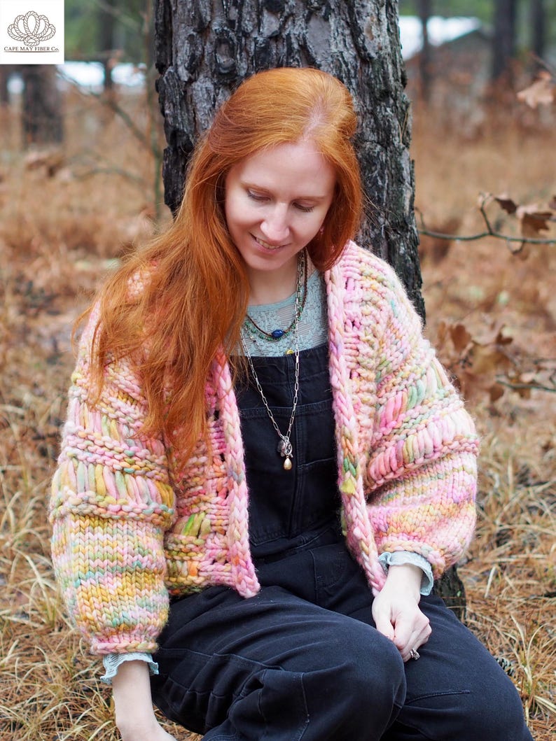 May include: A chunky knit cardigan in pastel shades of pink, yellow, and green. The open-front sweater has a boxy silhouette with wide sleeves. The garment is handmade with a textured stitch, and the sleeves are cuffed. The cardigan is worn over a dark jumpsuit.
