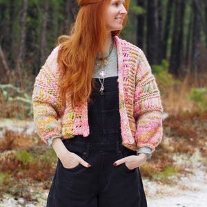 May include: A cropped, open-front cardigan in a mix of pastel colors, including pink, yellow, and green. The chunky knit sweater has a textured design and is worn over a light blue top and black overalls. The image is set in a forest.