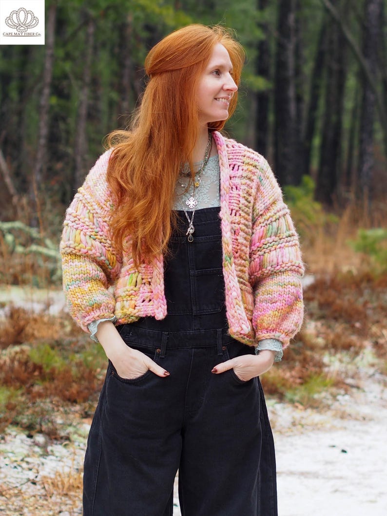 May include: A cropped, open-front cardigan in a mix of pastel colors, including pink, yellow, and green. The chunky knit sweater has a textured design and is worn over a light blue top and black overalls. The image is set in a forest.