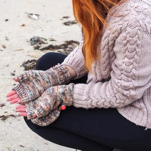 May include: A person wearing a light mauve cable-knit sweater and fingerless gloves. The gloves have a multicolored speckled pattern. The person is sitting on a sandy beach, wearing black pants.