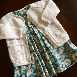 May include: White knitted cardigan with a button fastening and heart-shaped details on the pockets. The cardigan is lying on top of a blue and white floral dress.