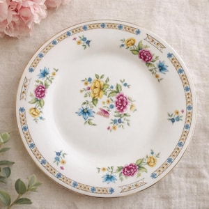 May include: White ceramic plate with a floral design featuring yellow, pink, and blue flowers. The plate has a gold rim and a decorative border with blue and gold accents. A classic piece for serving food.