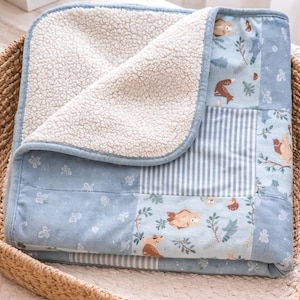 May include: A baby blanket with a patchwork design featuring woodland creatures and floral patterns in shades of blue, white, and brown. The blanket has a soft, white sherpa lining and a blue border. It is folded in a woven basket.