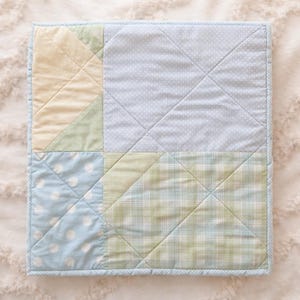 May include: A square patchwork quilt with a light blue border. The quilt features sections of light blue with white polka dots, yellow, green, and plaid patterns. Diagonal stitching creates a diamond pattern across the quilt.