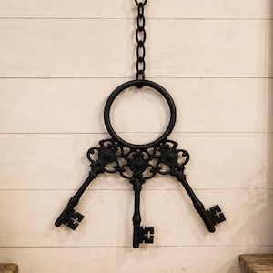 May include: A black metal wall hanging featuring three decorative keys suspended from a circular ring and a vertical chain. The keys are connected by an ornate, symmetrical metal filigree piece, creating a cohesive, singular wall decor item.
