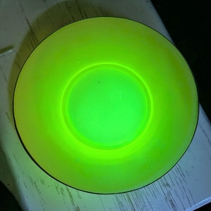 Può includere: Un piatto rotondo di vetro verde neon brillante. Il piatto ha un bordo leggermente rialzato e un centro incavato. Il colore è un verde vibrante, quasi elettrico, che suggerisce vetro all'uranio.