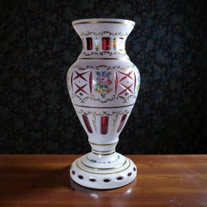 Pode incluir: Um vaso de vidro branco com detalhes em vermelho e dourado. O vaso apresenta um design floral e padrões recortados. A base e a borda são adornadas com ouro. O vaso tem cerca de 30 cm de altura.