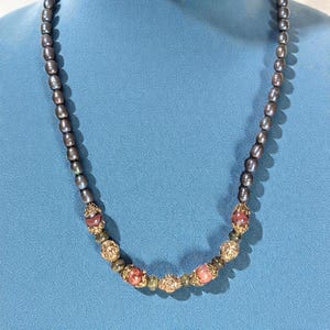 Puede incluir: Un collar con perlas grises oscuras y cuentas decorativas doradas. El collar incluye cuentas de acento rosas y doradas. El collar se muestra sobre un fondo azul.