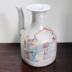 May include: A white ceramic pitcher with a long spout and a wide opening. The pitcher is decorated with a colorful scene of figures in traditional clothing. The scene includes figures in blue, pink, and orange clothing. The pitcher is on a dark wooden surface.