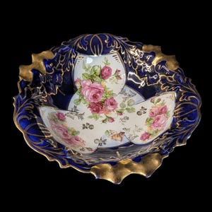 May include: A decorative three-part serving dish with a deep blue exterior and gold trim. The interior has a white background with pink roses and green leaves. The dish has a scalloped edge.