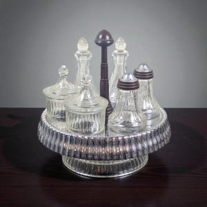 Könnte beinhalten: Ein Vintage-Gewürzset mit klaren Glasflaschen und -gläsern. Das Set umfasst Salzstreuer, Flaschen und Gläser mit Deckel, die auf einem mehrstufigen, kreisförmigen Tablett angeordnet sind. Ein zentraler, dunkelbrauner Griff ragt aus dem Tablett.