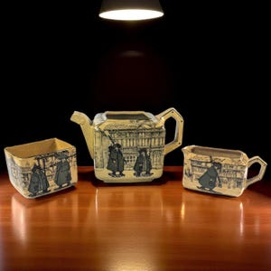 Puede incluir: Un juego de té de tres piezas que incluye una tetera, una jarra para la crema y un azucarero. Cada pieza está decorada con una ilustración en blanco y negro de figuras frente a un edificio. El juego tiene una combinación de colores crema y dorado.