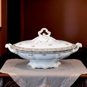 May include: A white porcelain serving dish with a lid, featuring a floral pattern in pink and green. The dish has a decorative handle and gold trim. It is placed on a table covered with a gray cloth.