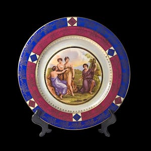 May include: A vintage porcelain plate with a decorative border in shades of blue and red. The center features a hand-painted scene of three women in a landscape setting.