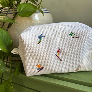 May include: White waffle-textured cosmetic bag with embroidered skiers in various colors. The bag has a zippered closure and a small loop on the side. The skiers are in green, blue, orange, and red. The bag is sitting on a green surface.