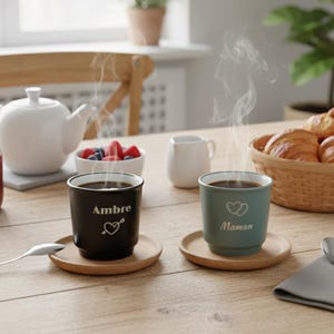 Op de afbeelding: Twee stomende koffiemokken op houten onderzetters. Eén zwarte mok heeft de naam "Ambre" met een hart- en pijldesign. De andere is blauwgroen met het woord "Maman" en een hartsymbool. Een theepot, room en gebak zijn op de achtergrond.
