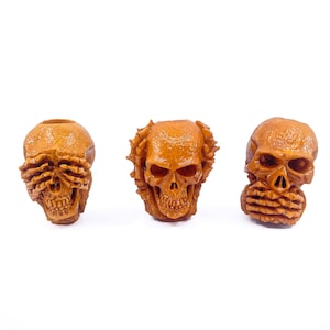 Puede incluir: Tres pequeñas figuritas de calavera de madera marrón. La primera calavera tiene los ojos cubiertos por manos, la segunda calavera tiene la boca cubierta por manos y la tercera calavera tiene las orejas cubiertas por manos.