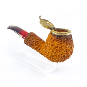Puede incluir: Una pipa de tabaco con un cuenco texturizado de color marrón claro y una boquilla marrón oscuro y roja. La pipa tiene una tapa dorada con bisagras y se exhibe sobre un soporte transparente. La pipa está diseñada para fumar.