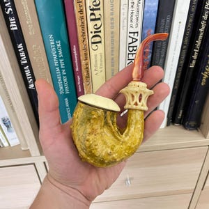 Puede incluir: Una pipa de tabaco amarilla y crema con un tallo curvo y una cazoleta decorativa tallada. La pipa tiene una tapa y una boquilla rojiza-anaranjada. La pipa se sostiene en una mano, con una estantería de fondo.