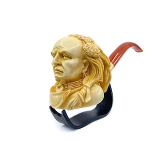 Può includere: Una pipa da tabacco scolpita con un busto dettagliato di un capo nativo americano. La pipa è color crema con un bocchino marrone rossiccio ed è esposta su un supporto nero. L'intaglio mostra dettagli intricati del volto e dell'acconciatura del capo.